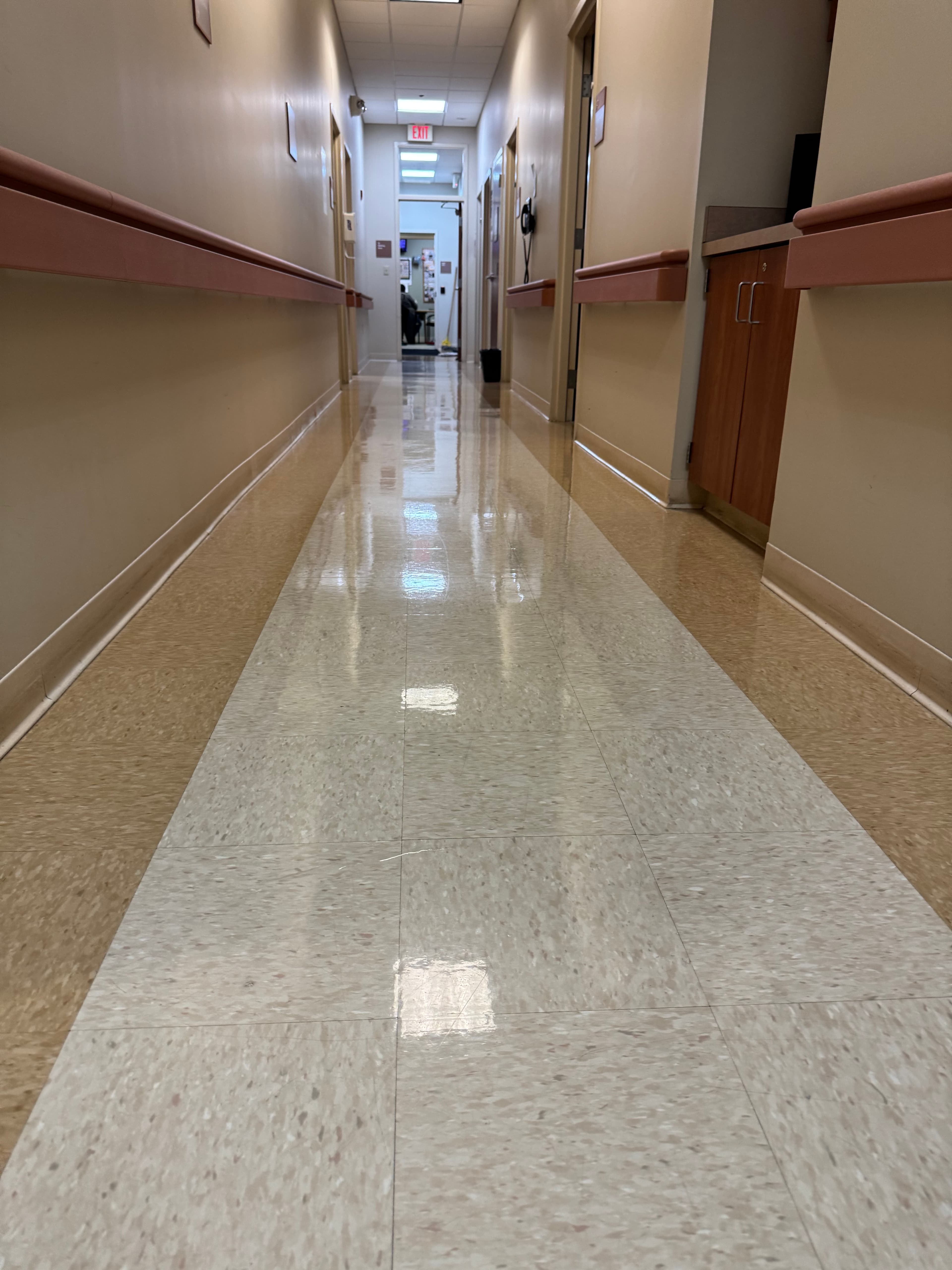 Long, empty hospital corridor featuring highly reflective floors, beige walls, and protective handrails.